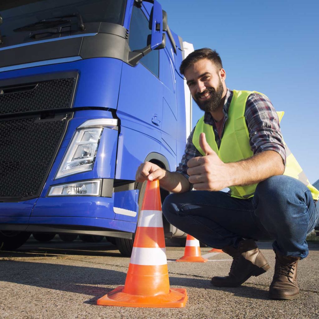 Shot of bearded man learning how to drive truck at driving schools. Truck driver candidate training for driving license. Standing by the traffic cone in reflective vest. Truck in the background.