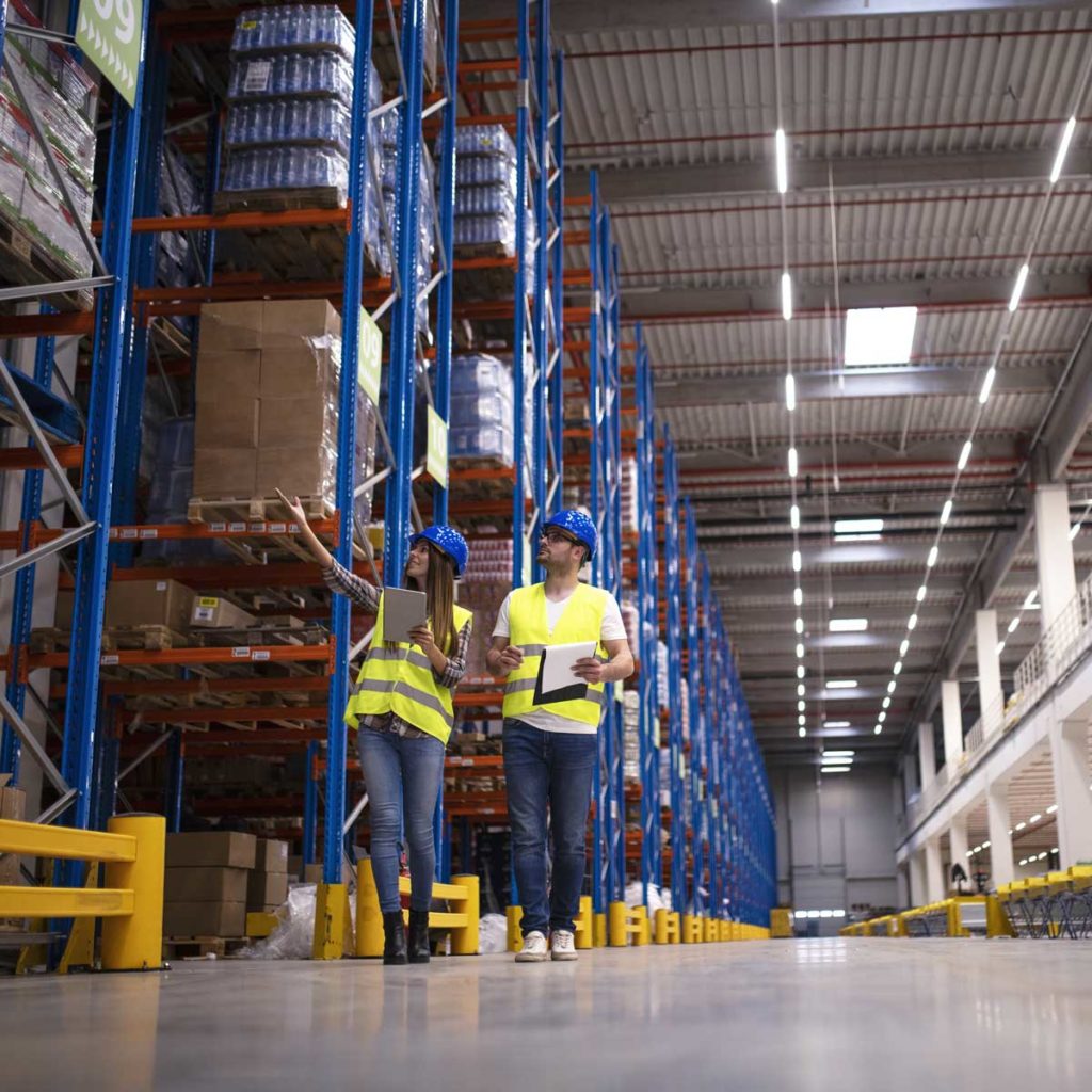 Shot of two workers walking through large warehouse center, observing racks with goods and planing distribution to the market. Storehouse department organization.