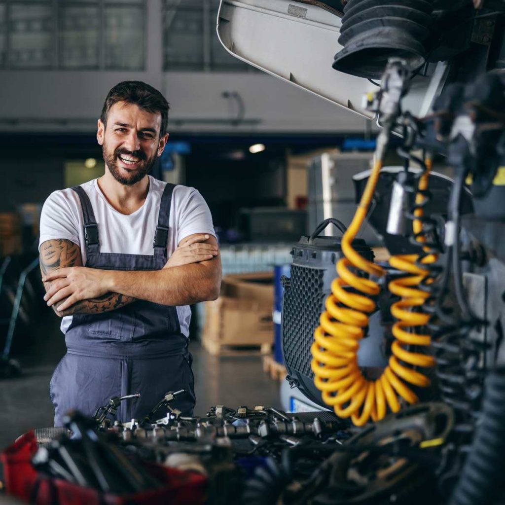 Smiling happy bearded tattooed worker in overalls standing next to truck with arms crossed.
